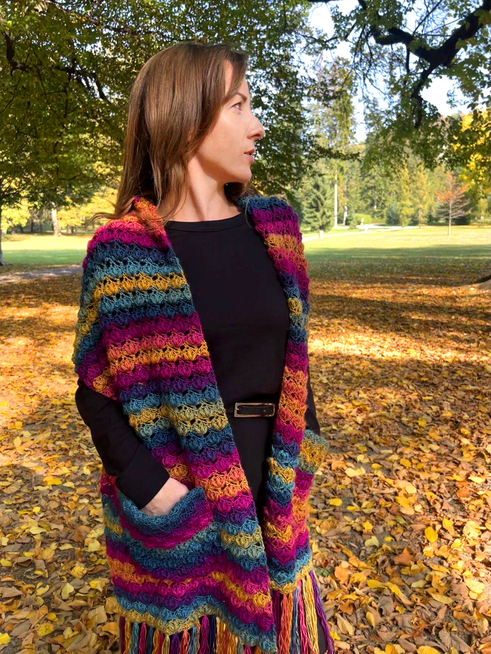 A woman standing outdoors in a park with fallen leaves, wearing a colorful striped crochet shawl over a black top.