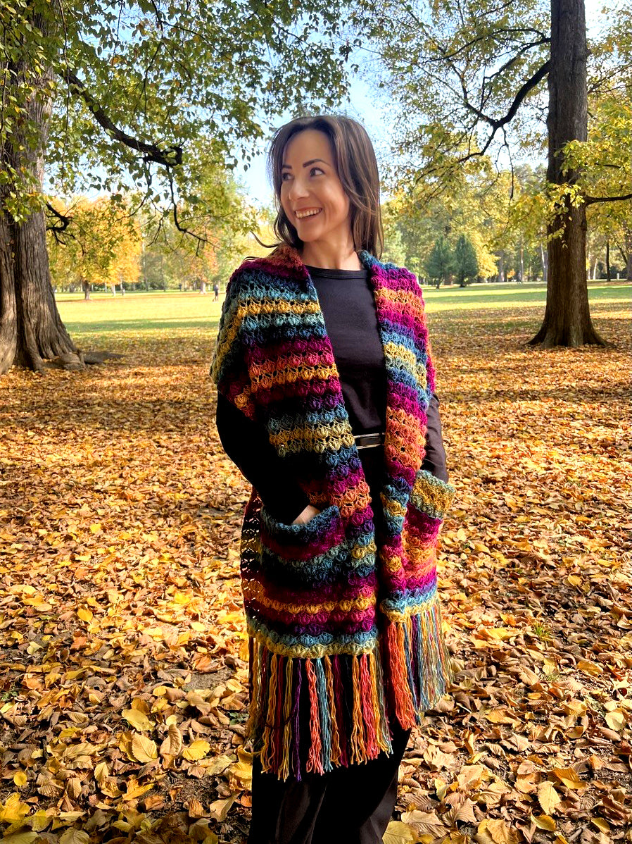 A woman standing in an autumn park wearing a vibrant, multicolored chunky crochet wrap with fringe, over a black top and black pants.