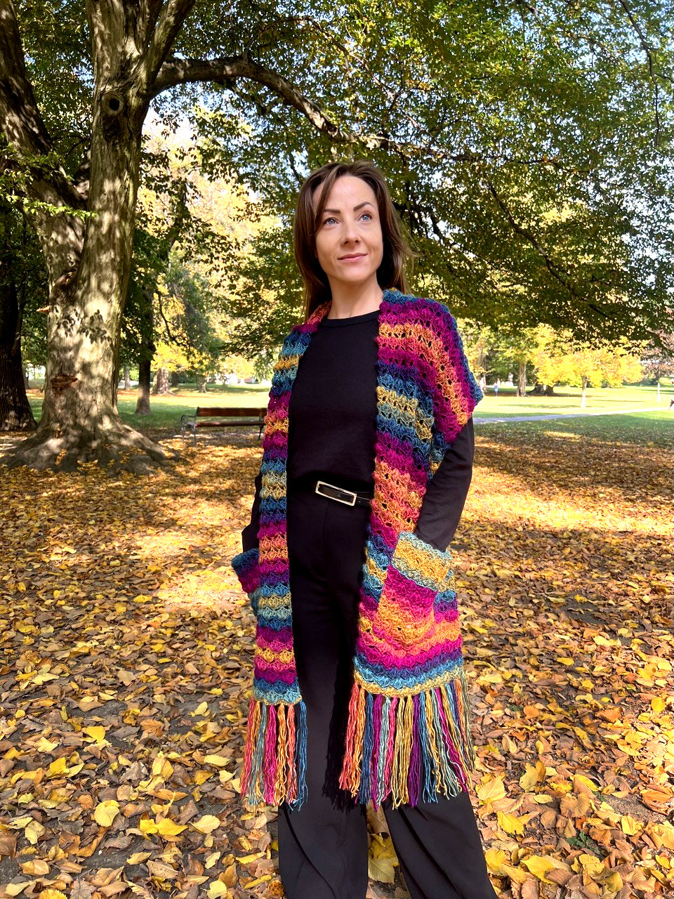 A woman standing in a park with fallen leaves, wearing a colorful striped crochet shawl with long fringe over a black outfit.