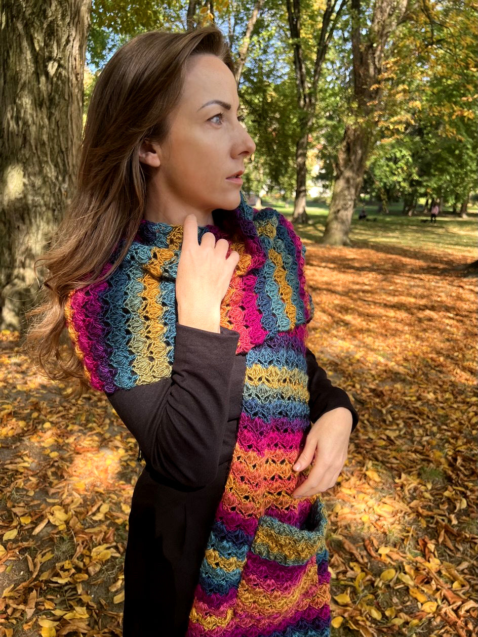 A woman wearing a vibrant, multicolored crocheted scarf with stripes of pink, blue, yellow, and orange, standing in an autumn park with fallen leaves.