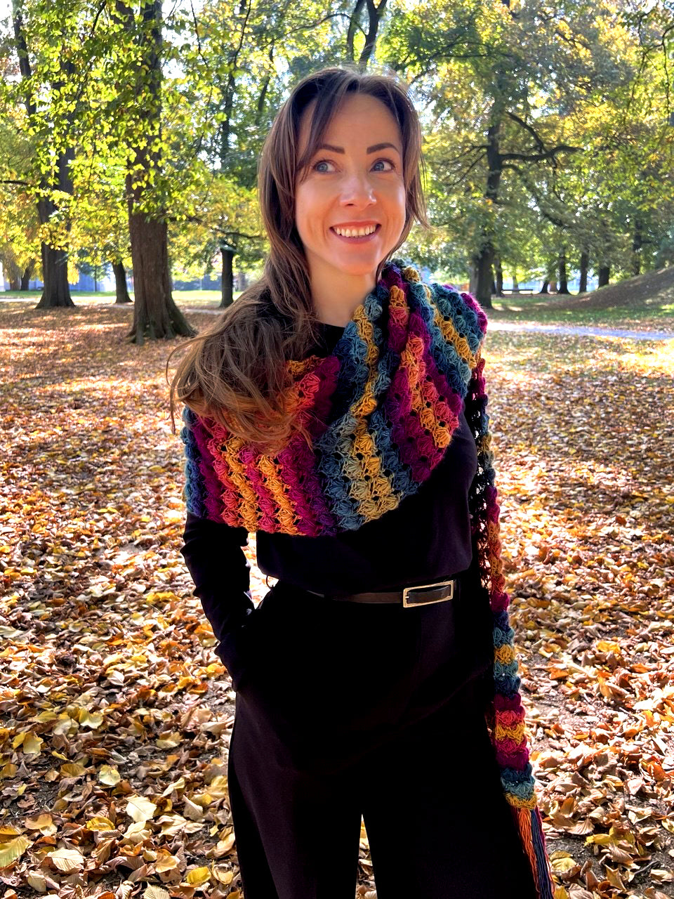 A woman standing in an autumn park wearing a thick, multicolored crocheted pocket scarf wrapped around her shoulders.