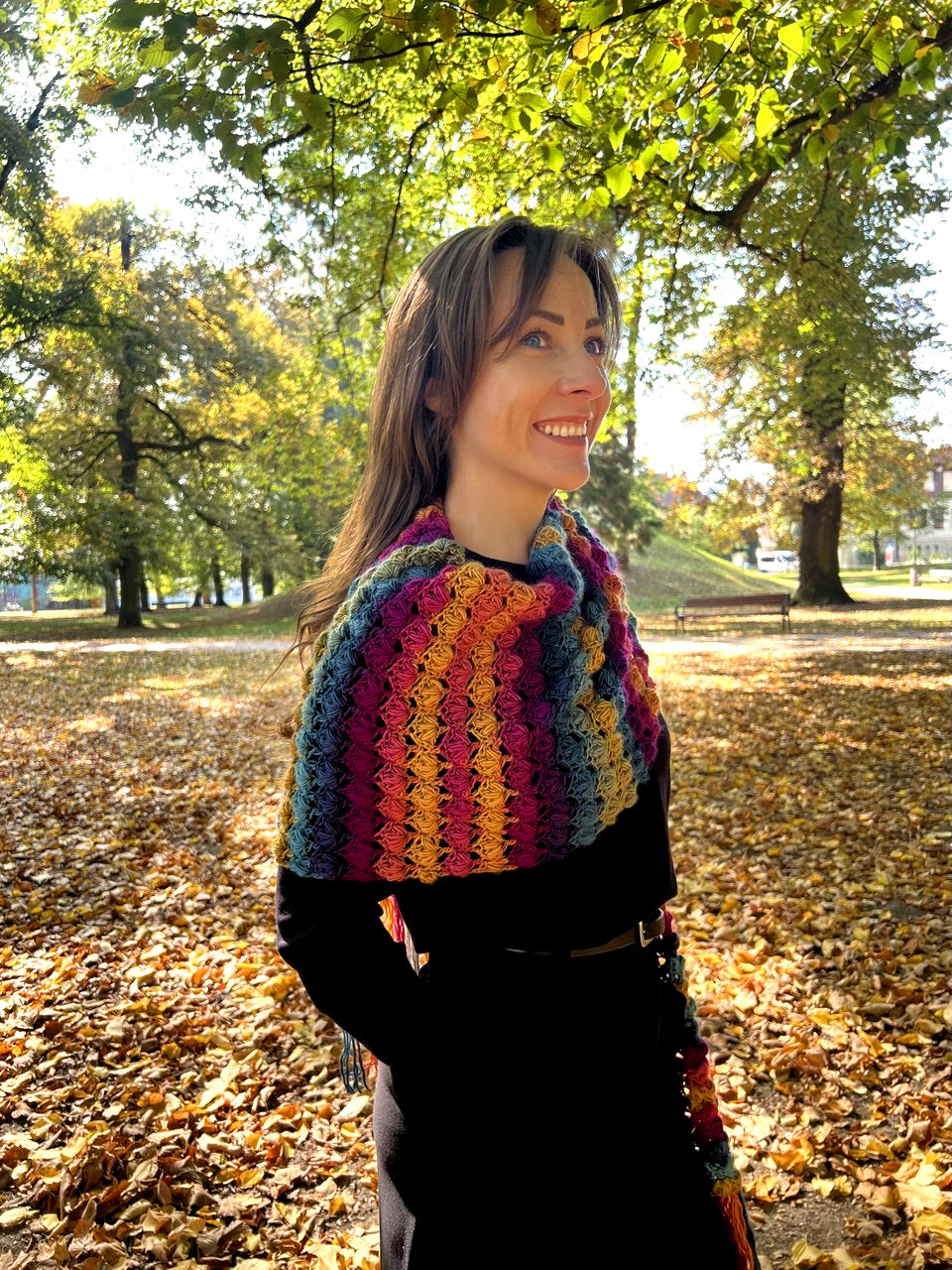 A woman wearing a vibrant, multicolored crocheted shawl or wrap over a black coat, standing in a park covered with fallen autumn leaves under leafy trees.