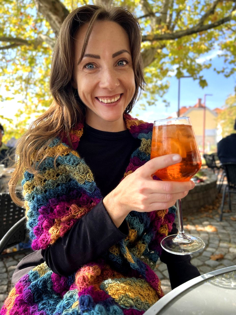 A smiling woman outdoors at a table holding a glass of Aperol Spritz, wearing a chunky multicolored crocheted Venezia pocket shawl over a black top.