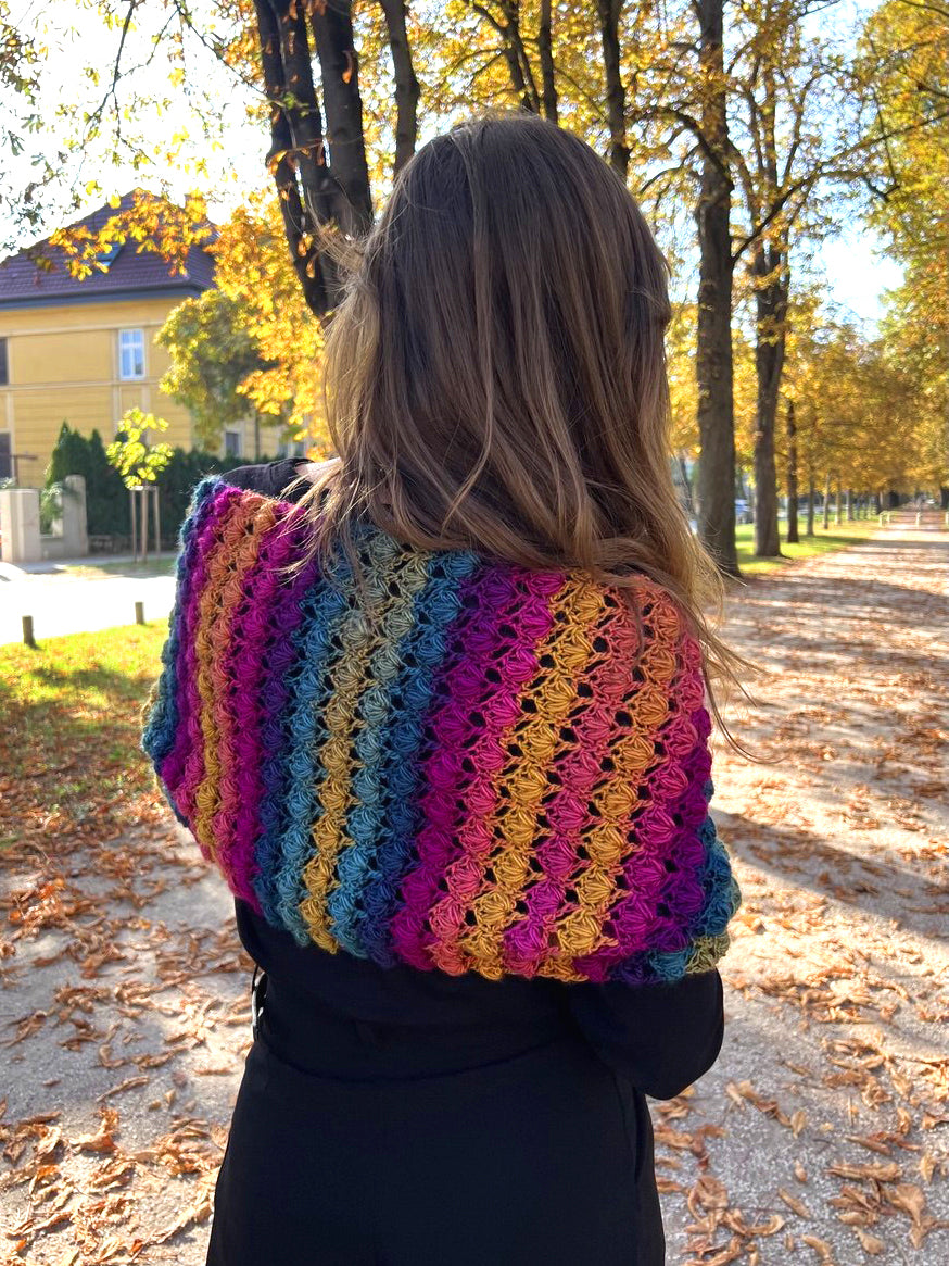 A woman viewed from behind wearing a vibrant, multicolored crocheted shawl or wrap with vertical wavy stripes in pink, purple, blue, yellow, and orange, set against an outdoor autumn park background with fallen leaves and trees.