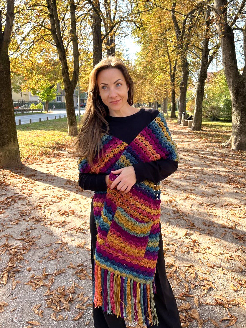 A woman standing in a park wearing a vibrant, multicolored striped knit wrap or shawl over a black long-sleeve top. The wrap features horizontal stripes of purple, blue, yellow, orange, and green with a fringed bottom.