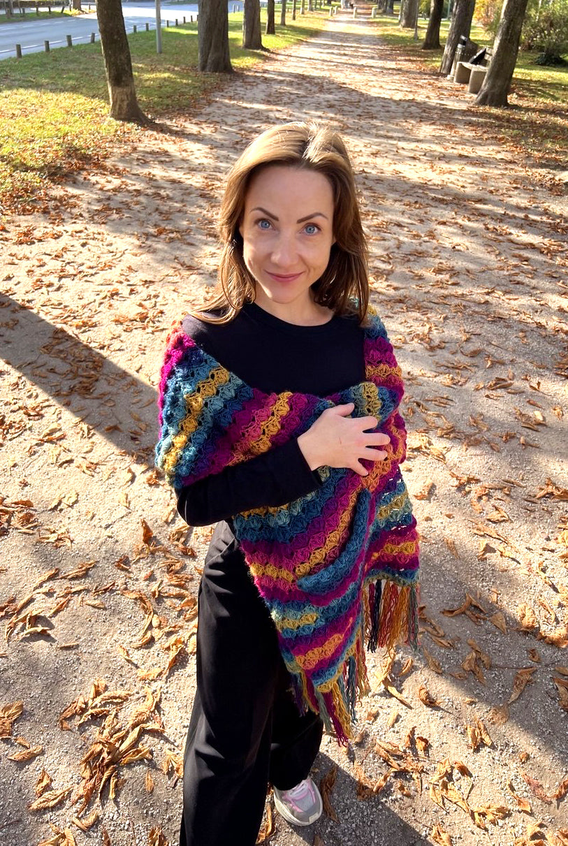 A woman standing on a leaf-covered outdoor path wearing a vibrant, multicolored striped crochet Venezia wrap over a black top and black pants.